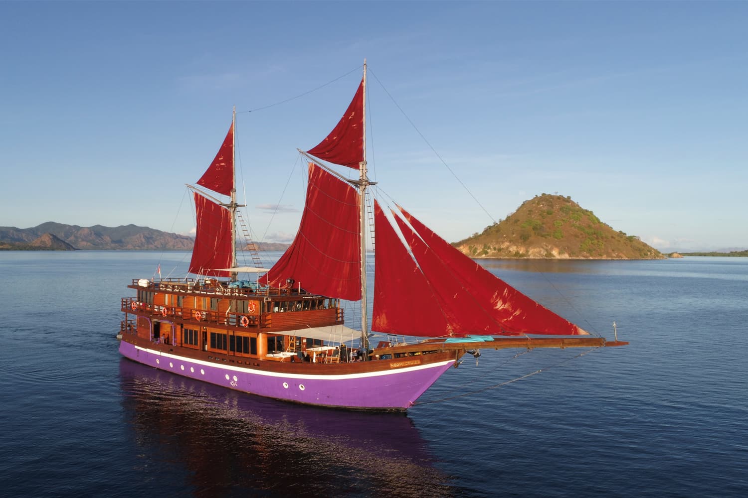 Neomi Cruise, a traditional Bugis phinisi liveaboard under full red sails off an Indonesian island